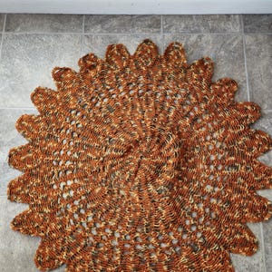 May include: A large, round, crocheted rug in shades of brown, beige, and gold. The rug has a lacy, openwork design and a scalloped edge.