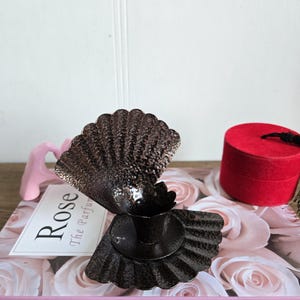 Puede incluir: Un portavelas decorativo de metal marrón oscuro con diseño de concha. El portavelas está sobre un libro con las palabras "Pink Rose" y "Rose The Parfum". También se ve una pequeña caja roja.