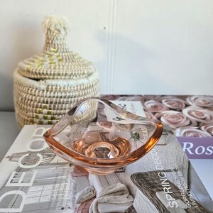 Op de afbeelding: Een kleine, roze glazen kom met een unieke, gebogen vorm. De kom staat op een boek met de woorden "Home Decor" en "Spring Decorating Ideas". Een geweven mand met een deksel staat op de achtergrond.