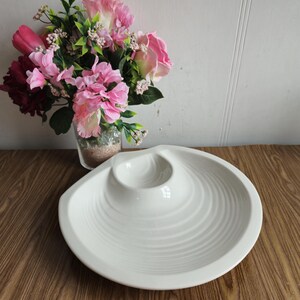 Puede incluir: Plato de servir de cerámica blanca con una sección central elevada, que se asemeja a un volcán o una concha. El plato tiene una textura ondulada y es perfecto para servir salsas, aperitivos o entrantes.