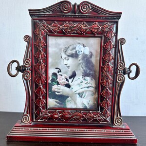 Op de afbeelding: Een rood houten fotolijst met sierlijke details en een sleutelgat aan elke kant. De lijst bevat een zwart-witfoto van een jonge vrouw met bloemen.