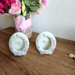 Peut inclure: Deux petits cadres photo ovales en porcelaine avec un motif floral dans des tons de rose, bleu et vert. Les cadres sont posés sur une surface en bois.