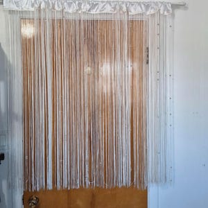 Könnte beinhalten: Weißer Perlenvorhang, der vor einer Holztür hängt. Der Vorhang hat eine weiße Satin-Kopfleiste und besteht aus zahlreichen vertikalen Strängen mit kleinen, silbernen Perlen. Die Tür ist braun mit einem Messing-Türknauf.