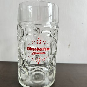 May include: A clear glass beer stein with a red and white Oktoberfest Rickards' logo. The stein has a traditional German design with a handle and a lid.