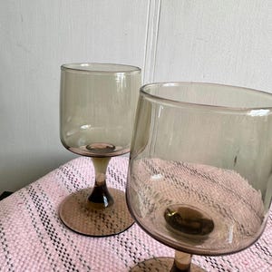 May include: Two clear glass wine glasses with brown tinted bases. The glasses are on a pink and white striped surface.