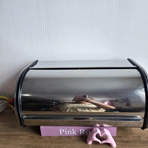 Könnte beinhalten: Ein Brotbehälter aus Edelstahl mit schwarzem Rand. Er steht auf einer Holzoberfläche, mit einem rosa Schild mit der Aufschrift "Pink Ro" und einer rosa Hand-Herz-Skulptur.