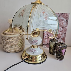 Peut inclure: Une lampe de table vintage avec une base et des accents dorés. La lampe présente un corps en céramique blanche orné de motifs floraux roses et un abat-jour en verre avec un motif floral. Un panier en osier et des bougeoirs décoratifs sont également visibles.