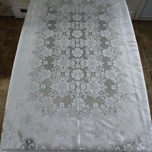 Peut inclure: Nappe en dentelle blanche avec un motif floral. La nappe rectangulaire recouvre une table. Le motif en dentelle présente des motifs détaillés de fleurs et de feuilles. Les bords de la nappe ont une bordure festonnée.