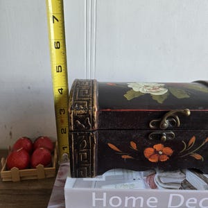May include: A black wooden chest with floral designs in white and orange, measuring approximately 4 inches in height. The chest has a decorative clasp and is placed on top of books. The chest is a decorative storage item.