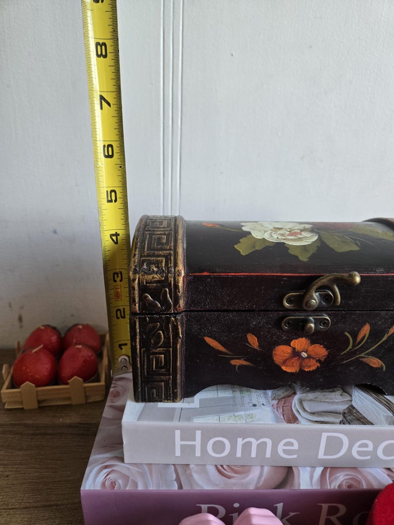 May include: A black wooden chest with floral designs in white and orange, measuring approximately 4 inches in height. The chest has a decorative clasp and is placed on top of books. The chest is a decorative storage item.