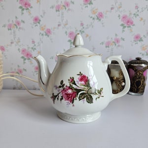May include: A white porcelain teapot featuring a floral pattern of pink roses and green leaves on the body. The teapot has a matching lid with a pointed finial, a curved handle, a spout, and gold trim along the scalloped rim and edges.