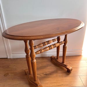 May include: A small, oval-shaped wooden table with a simple design. The table has a light brown finish and features turned legs with a decorative base.