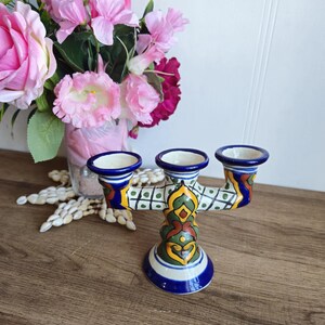 May include: A three-armed ceramic candle holder with a blue, green, yellow and red painted design. The candle holder is on a wooden surface.