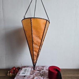 Peut inclure: Une lanterne suspendue orange en forme de cône avec une armature et une chaîne en métal noir. La lanterne est suspendue au-dessus d'une boîte de roses, d'un petit fez rouge et d'une sculpture rose en forme de main. Le texte "Rose The Perfumers" est visible.