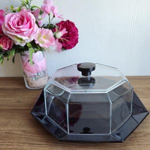 May include: Octagonal glass cake dome with a black base and a black knob on top. The dome is sitting on a wooden surface.