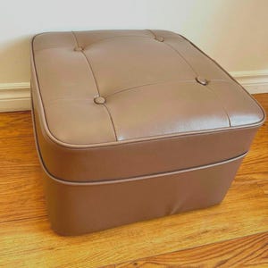 Puede incluir: Un otomano de cuero marrón con una parte superior capitoné y una forma cuadrada.