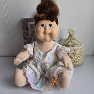 Könnte beinhalten: Eine Vintage-Puppe mit braunen Haaren im Dutt, blauen Augen und einem lächelnden Gesicht. Die Puppe trägt ein weißes Kleid mit bunten vertikalen Streifen und rosa Besatz. Die Puppe sitzt auf einer weißen Oberfläche.