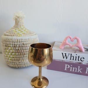 Puede incluir: Una copa de vino dorada con un cuenco redondeado y un tallo delgado. La copa se exhibe sobre una superficie blanca, junto a una cesta tejida con tapa y una escultura rosa en forma de corazón. El fondo incluye libros con las palabras "White" y "Pink".