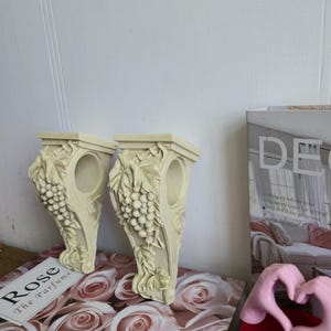 Peut inclure: Deux appliques murales décoratives blanc cassé avec des détails de grappes de raisin et de feuilles. Les appliques sont posées sur une surface à motif de roses. Un magazine et une pancarte "Rose The parfume" sont également visibles.