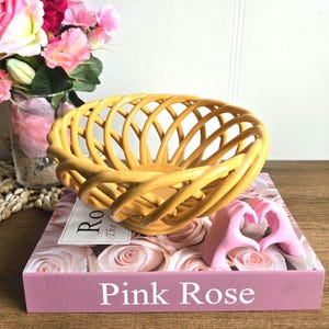 Peut inclure: Un bol en céramique jaune tressé posé sur un livre rose avec le titre "Pink Rose" et une sculpture de main rose formant un cœur.