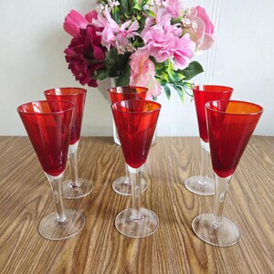 Peut inclure: Ensemble de six verres à shot en verre rouge avec des tiges claires. Les verres sont disposés en demi-cercle sur une surface en bois.