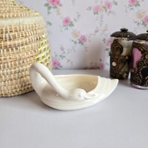 May include: A white ceramic swan-shaped dish with a smooth, glossy finish. The swan's neck curves gracefully to form the handle, while the body of the bird serves as the shallow bowl. The design features subtle horizontal ridges along the tail.