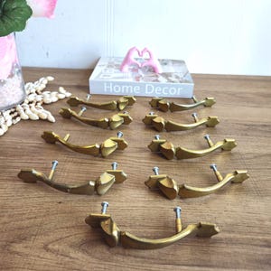 以下が含まれることがあります： 装飾的な華やかなデザインの金色のキャビネットノブが10個。ノブは湾曲した形をしていて、先端が尖っています。ノブは木製の表面に5個ずつ2列に並んでいます。