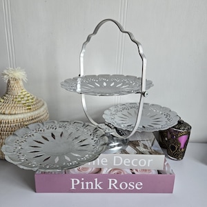 May include: A silver, three-tiered serving tray with a handle. The tray has a decorative, openwork design. The base of the tray is resting on a stack of books with the text "Pink Rose" visible. A decorative basket and a small purple container are also in the image.