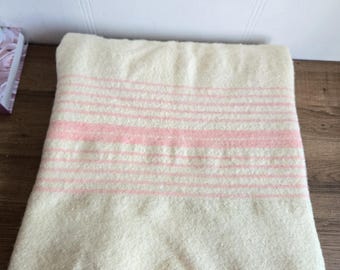 Puede incluir: Una manta plegada, de color crema, con rayas horizontales en un tono rosa suave. La manta está hecha de un material grueso y texturizado, y las rayas están espaciadas uniformemente a lo ancho. La manta está cuidadosamente doblada.