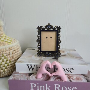 Peut inclure: Un cadre photo orné en bronze foncé avec un intérieur bleu marine est posé sur deux livres. Les livres sont intitulés "White Rose" et "Pink Rose". Une paire de mains roses forme un cœur. Un panier tressé est en arrière-plan.