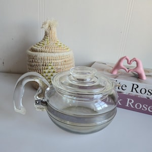 Peut inclure: Une théière en verre transparent avec un couvercle et une poignée. La théière a une bande argentée au milieu. Un panier tressé avec un couvercle et une sculpture rose en forme de main sont en arrière-plan.