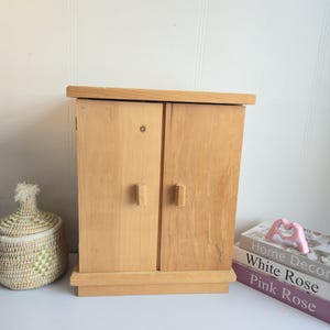 Puede incluir: Un pequeño armario de madera clara con dos puertas y tiradores rectangulares. El armario se asienta sobre una base y tiene un diseño sencillo. Una cesta decorativa y libros están al lado del armario.