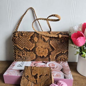 May include: A snakeskin-patterned handbag with a braided strap and a small matching coin purse. The bag and purse are brown and tan with a scale pattern. The bag is rectangular with a flap closure.