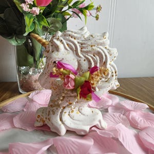Peut inclure: Figurines de licorne en céramique blanche avec des accents de paillettes dorées et une couronne de fleurs roses. La licorne est assise sur un lit de pétales de roses roses.