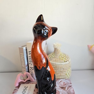 May include: A ceramic cat figurine with a glossy finish. The cat is primarily black with orange and brown accents. The figurine is posed in a seated position with its head turned. The cat is placed on a surface with a floral pattern.