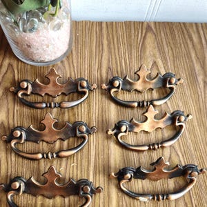 Puede incluir: Seis tiradores de armario de estilo antiguo con acabado en cobre y diseño ornamentado. Los tiradores tienen una base de metal negro y una parte superior decorativa en forma de corona.