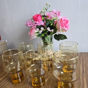 Può includere: Set di sei bicchieri in vetro ambrato con una base larga. I bicchieri sono trasparenti e hanno un aspetto vintage.