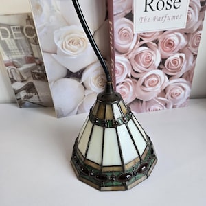 Peut inclure: Une petite lampe à suspension en vitrail avec un motif géométrique blanc et vert. La lampe a une base en métal de couleur bronze et un cordon noir. L'arrière-plan comprend un livre intitulé "Rose" et d'autres objets décoratifs.