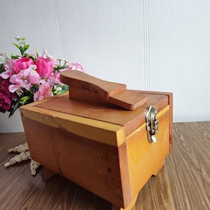 Könnte beinhalten: Eine Schuhputzkiste aus Holz mit einem Scharnierdeckel und einem kleinen Holzschuhspanner oben. Die Kiste hat einen Riegel und ist aus hellem braunem Holz.