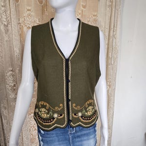 Peut inclure: Gilet vert olive avec bordure noire et broderie dorée. Le gilet a un col en V et une broderie florale décorative en bas. Le gilet est sans manches et possède des boutons noirs sur le devant.