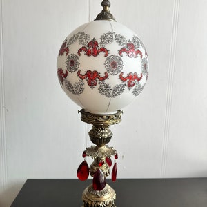 Peut inclure: Une lampe globe blanche avec un motif floral noir et rouge. La lampe a une base en métal doré avec des accents de cristal rouge.