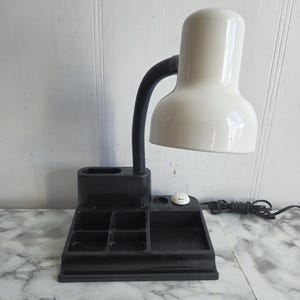 May include: A black desk lamp with a white shade and a built-in organizer with multiple compartments. The lamp has a flexible neck and a cord.