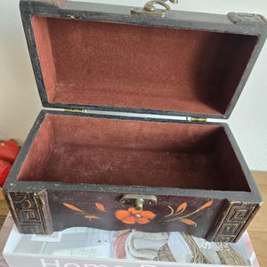 May include: Open, antique-style wooden chest with a dark brown exterior and a red velvet interior. The chest features a floral design with orange flowers and gold accents. The chest is resting on a stack of books with the text "Home Decor" visible.