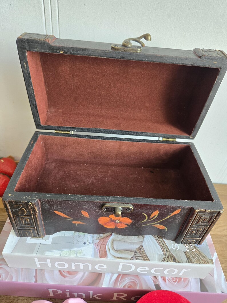 May include: Open, antique-style wooden chest with a dark brown exterior and a red velvet interior. The chest features a floral design with orange flowers and gold accents. The chest is resting on a stack of books with the text "Home Decor" visible.