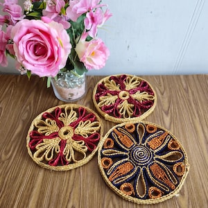Peut inclure: Trois dessous-de-plat en paille tressée avec des motifs colorés. Les dessous-de-plat sont ronds et présentent un motif floral. Un dessous-de-plat est rouge et or, un autre est bleu et orange, et le dernier est or et brun.