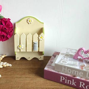 May include: A yellow wooden key holder with a picket fence design and a small milk can. The holder has three hooks and is decorated with white daisies. The key holder is sitting on a wooden surface with a pink book and a pink heart-shaped object.