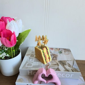 Puede incluir: Un palillero decorativo con forma de sándwich con palillos de sándwich en miniatura. El soporte es amarillo con detalles rojos y verdes, y se encuentra sobre una pila de libros. Una escultura de manos rosas forma un corazón.