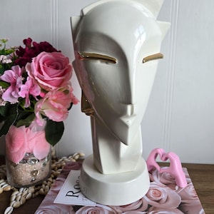 May include: White ceramic sculpture of a stylised face with gold accents. The sculpture has a geometric design and is a modern art piece.