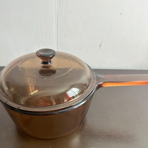 Puede incluir: Una cacerola de vidrio marrón con tapa y asa. La cacerola está sobre una superficie marrón oscura.