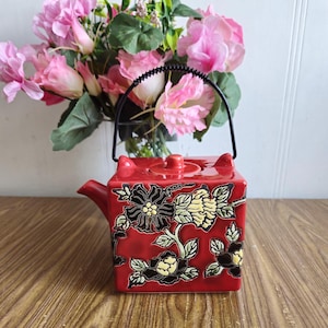 Puede incluir: Tetera de cerámica roja con asa de metal negro y diseño floral. La tetera tiene un cuerpo cuadrado y una tapa con un pomo.